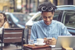 A young man is sitting at a sidewalk cafe and smiling at the phone he's holding in both hands. He's wearing a light blue, button-down shirt and has large, stylish glasses pushed up on his head.